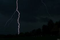 Lightning bolt striking from dark cloudy sky over trees and a building at night.