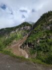 Mountain landscape with green vegetation, rocky slopes, narrow waterfall, dirt path, and cloudy sky.