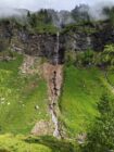 Waterfall flows down rocky cliff through green hillside with mist and scattered trees.