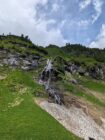 Mountain slope with green grass, sparse trees, waterfall, melting snow patch, and partly cloudy sky.