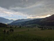 Valley with green fields and scattered trees, a small town, forested hills, and sun rays through clouds.