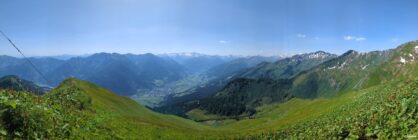 Panoramic view of green mountain slopes, a valley town, and snow-capped peaks under blue sky
