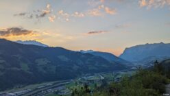 Mountain valley with scattered buildings, green fields, a winding river, and sunset sky.