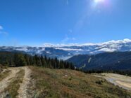 Mountain trail with pine trees, lake, and cloud-covered mountains under a sunny blue sky.