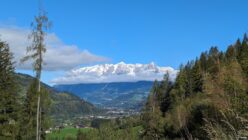 Green valley with houses, forested hills, and snow-capped mountains partially covered by clouds under blue sky.