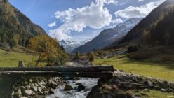 Wooden bridge over stream in mountain valley with autumn trees and snow-capped peaks.