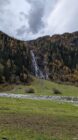 Grassy meadow, rocky slope with waterfall, and forested hillside with autumn foliage under cloudy sky