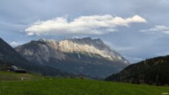 Green field bordered by forested hills with a rocky mountain range and partly cloudy sky above.